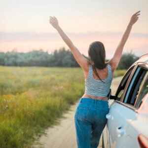 A woman stands beside a car on a summer field road, arms raised in freedom, enjoying a beautiful sunset.